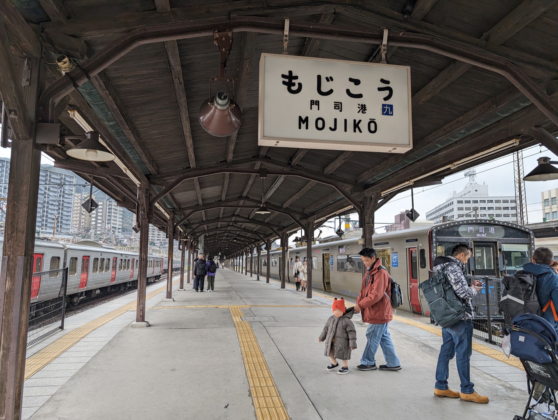 View along a long railway station platform covered the a wooden canopy, with silver metal and red trains on both sides of the platform